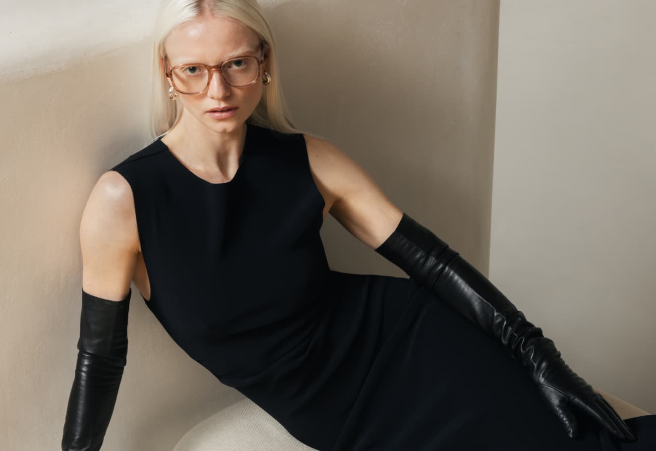 Ace & Tate | A blonde woman modeling translucent glasses and long black leather gloves while reclining against a curved beige wall.