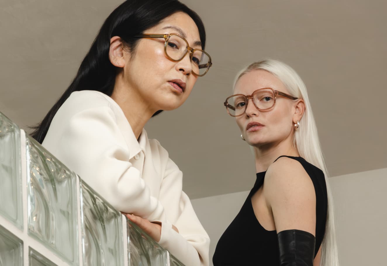 Ace & Tate | Two women modeling eyewear near a glass-block wall; one wears a black top with leather gloves, the other a cream top.