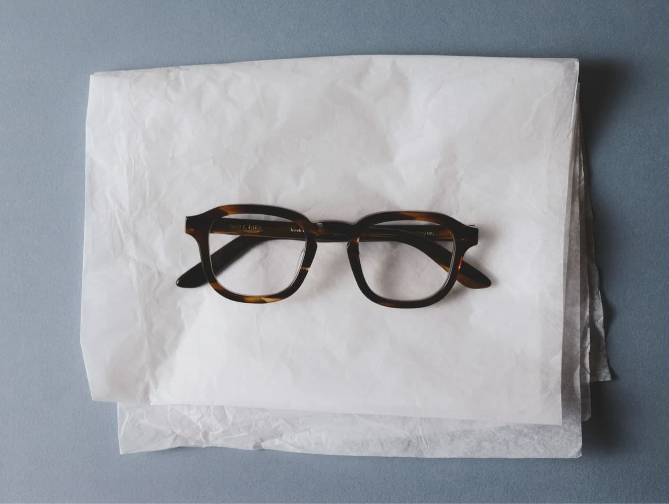 Ace & Tate | A pair of thin, round metal-framed glasses nesting in crumpled craft paper inside a small cardboard box.