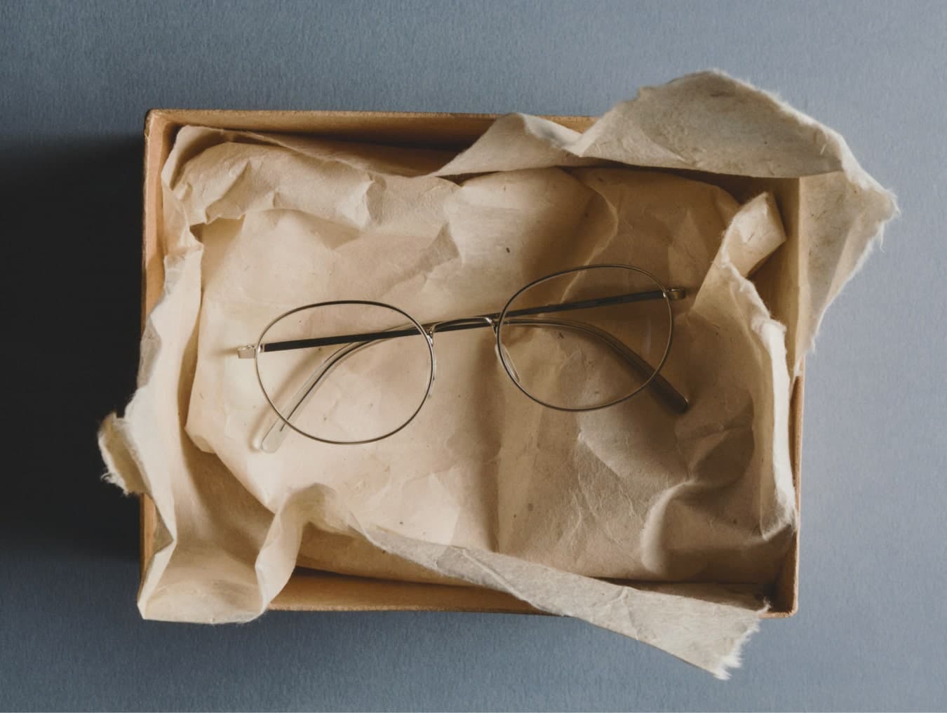 Ace & Tate | A pair of thick-rimmed tortoiseshell eyeglasses rests centrally on a stack of folded white paper against a muted blue background.